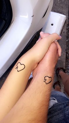 two people holding hands with tattoos on their arms and the other hand is in front of a white car