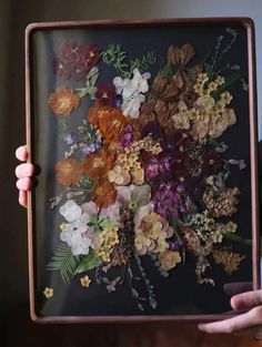 a person holding up a framed painting with flowers on it's side and leaves in the middle