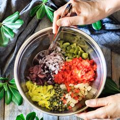 hands mixing vegetables in a bowl on top of a wooden table next to green leaves