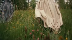 two women in dresses are walking through tall grass and flowers with trees in the background