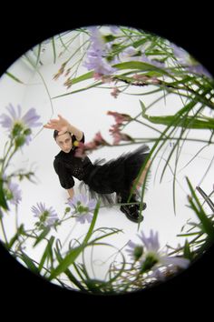 a small doll is posed in front of some purple flowers and green leaves on a white surface