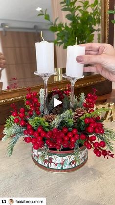 someone is holding two candles in front of a christmas centerpiece with holly and berries