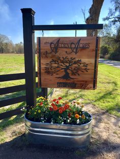 a wooden sign sitting on top of a metal planter filled with flowers next to a tree