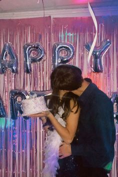 a man and woman kissing in front of a birthday cake with balloons on the wall behind them