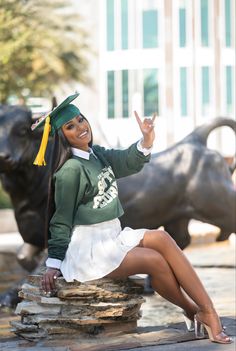 a woman in a green and white graduation outfit is sitting on a rock with her hand up