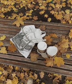 a newspaper and cups on a wooden bench in the fall leaves with someone's feet