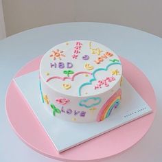 a white cake with rainbows and clouds on it sitting on a pink plate next to a book