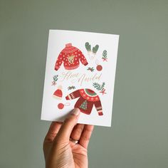 a hand holding up a card with an ugly sweater on it and christmas decorations around it