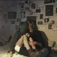 two people sitting on a bed kissing in front of a wall covered with pictures and post it notes