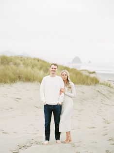 an engaged couple standing on the beach in front of some tall grass and sand dunes