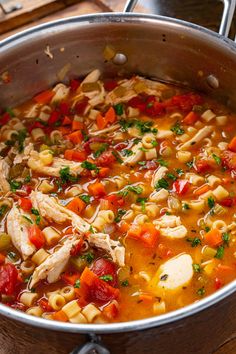 a pot filled with pasta and chicken soup on top of a wooden table next to utensils