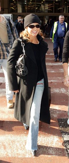 a woman in black coat and jeans walking down the street with a handbag on her hip