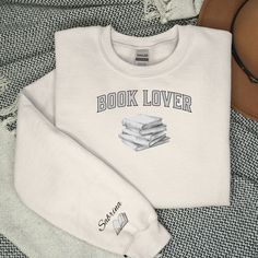 This personalized hoodie or sweatshirt is for the person in your life that loves books and reading.  The design features a university style font across the front that reads "Book Lover" with a graphic of a stack of books below it.  The hoodie (or sweatshirt) is personalized by adding the recipient's name to the outside of the right sleeve with an open book printed below it.  A book lover will enjoy this shirt anytime, but keep it in mind as a gift idea for birthdays, Christmas, etc.   This unise White Bookish Sweatshirt With Letter Print, Book Sweatshirts, Aesthetic Hoodies, Purple Books, University Style, Personalized Hoodies, Open Book, Stack Of Books, Sweatshirt Shirt