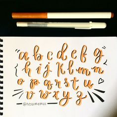 the letters are drawn in orange ink on a white paper with two pencils next to it