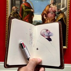 a person holding an open book in front of a painting with a pen on it