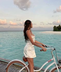 a woman in a white dress standing next to a bike near the water with an island in the background