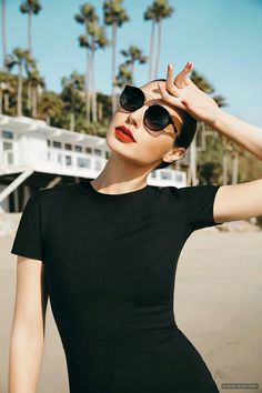 a woman in black dress and sunglasses standing on the beach with palm trees behind her