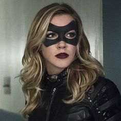 a woman wearing a black mask and leather jacket with long blonde hair is looking at the camera