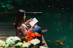 a woman sitting on a bench reading a book near a pond with goldfish in it