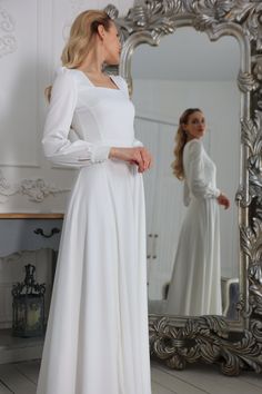 a woman standing in front of a mirror wearing a white dress with long sleeves and an open back