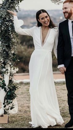 a bride and groom holding hands under an arch with greenery on the side, smiling at each other