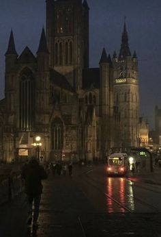 people are walking on the sidewalk in front of an old church at night with traffic passing by