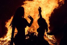 two women standing in front of a fire with their hands up to the sky,