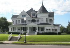 a large white victorian style house sitting on the side of a road next to a lush green field