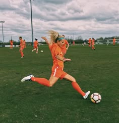 a woman in an orange uniform kicking a soccer ball on a field with other players