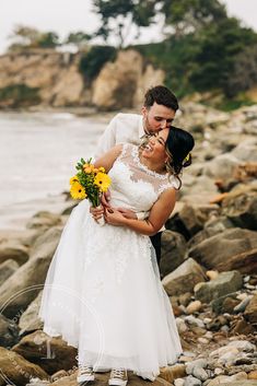 a bride and groom standing on rocks by the water with their arms around each other