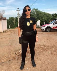 a woman standing in the dirt wearing sunglasses and a black shirt with gold medallions