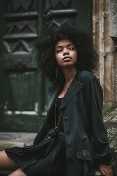 a woman with an afro sitting on the ground in front of a green door wearing a black coat