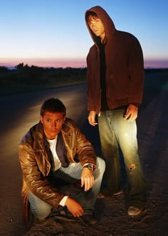 two men sitting on the side of a road at night, one is wearing a brown jacket