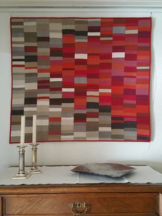 a red and grey quilt hanging on the wall next to a table with two candles