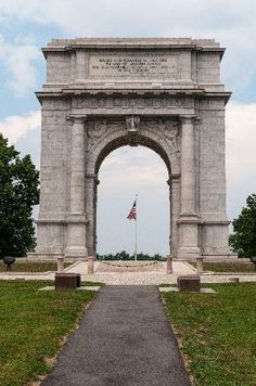 an arch in the middle of a grassy area with a flag on top and trees around it