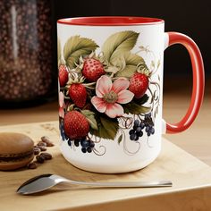 a coffee mug with strawberries and flowers painted on the side next to a spoon