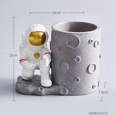 an astronaut figurine next to a silver cup on a white surface with measurements