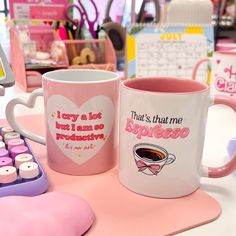 two coffee mugs sitting next to each other on a pink table with keyboard and mouse