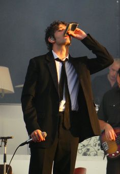 a man in a suit and tie drinking from a cup while standing next to a guitar