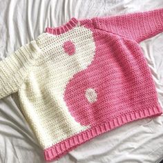 a pink and white knitted sweater laying on top of a bed next to a pillow