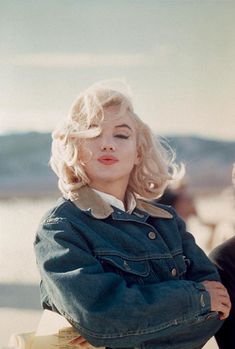 a woman with blonde hair wearing a jean jacket and sitting on a bench near the ocean