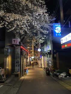 a narrow city street at night with white flowers on the trees and people walking down the sidewalk