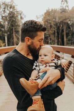 a man holding a baby in his arms on a bridge over looking the water with trees in the background