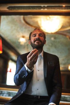 a man in a tuxedo is sitting on a chair with his hands clasped