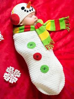 a baby wearing a knitted snowman hat and scarf laying on a red blanket