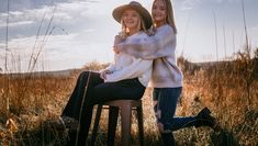 two young women sitting on stools in tall grass, one with her arms around the other