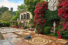 a large clock mounted to the side of a wall covered in plants and flowers next to a walkway