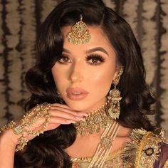 a woman with dark hair wearing gold jewelry and makeup looks at the camera while holding her hand near her face