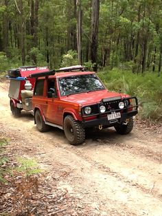 a red jeep driving down a dirt road in the woods with a trailer attached to it's back