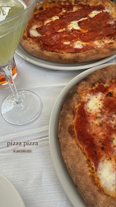 two pizzas sitting on top of white plates next to a glass of wine and a drink